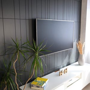 May include: A modern living room setup featuring a large, flat-screen television with a silver frame mounted on a dark gray paneled wall. A white cabinet below holds decorative items, including a vase with dried plants, books, and wooden letters spelling "HOME". Green plants add a natural touch.