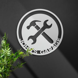 Könnte beinhalten: Ein rundes Schild aus weißem Metall mit einem Schraubenschlüssel und einem Hammer, die sich kreuzen. Das Schild ist rund und hat den Text "John's Garage" am Rand.