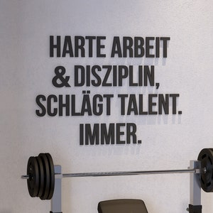 May include: A weight bench with a barbell and weights. The wall behind the bench has a motivational quote in German: "Harte Arbeit & Disziplin, schlägt Talent. Immer."