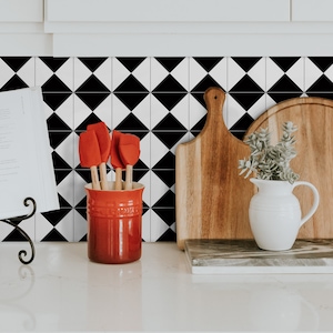May include: A red ceramic utensil holder with a set of red silicone spatulas on a white worktop with black and white diamond patterned tile splashback. A wooden chopping board and a white jug with a green plant are also on the counter.