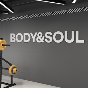 Puede incluir: Interior de gimnasio con las palabras "BODY & SOUL" en letras blancas sobre una pared gris. Se ve un soporte de pesas con pesas amarillas. El techo tiene detalles a rayas blancas y negras e iluminación cenital.
