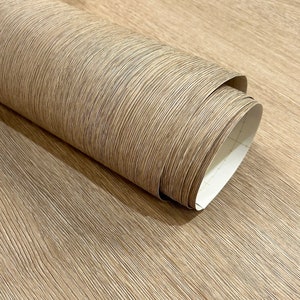 May include: A roll of light brown wood-look wallpaper with a textured, linear pattern. The wallpaper is partially unrolled, revealing the white backing. The wood grain design adds a natural aesthetic.