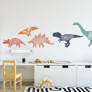 Könnte beinhalten: Eine weiße Wand mit fünf Dinosauriersilhouetten in verschiedenen Farben. Die Dinosaurier sind mit ihren Namen beschriftet: Pterodactylus, Stegosaurus, Tyrannosaurus, Triceratops und Apatosaurus.