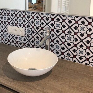 May include: A white ceramic vessel sink sits on a wood-grain vanity countertop. Behind the sink, a wall features square tiles with a repeating black, white, and maroon heart-shaped pattern and blue star accents. A chrome faucet and electrical outlets are visible.