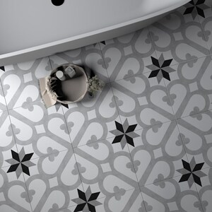 May include: A white bathtub with a black drain sits on a floor covered in square tiles featuring a repeating pattern of gray heart-like shapes and black eight-pointed stars. A small round basket containing white items sits on the tiled floor.