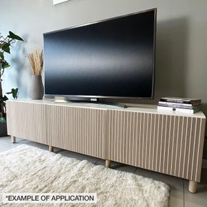 May include: A modern, light-colored TV stand with vertical wood grain detailing. The stand features a white top, light brown doors, and short, rounded wooden legs. A large flat-screen TV sits atop the stand, with decorative items on the surface.
