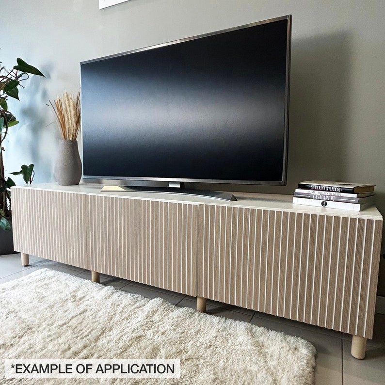 May include: A modern, light-colored TV stand with vertical wood grain detailing. The stand features a white top, light brown doors, and short, rounded wooden legs. A large flat-screen TV sits atop the stand, with decorative items on the surface.