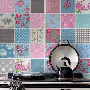 May include: A black stovetop kettle and a metal lid resting on a stove against a wall covered in square tiles featuring various patterns including pink roses, blue swirls, white polka dots, grey checks, and heart shapes in blue and pink.
