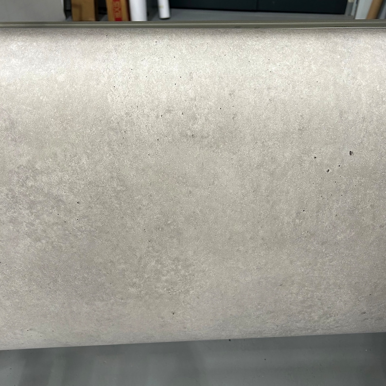 K&ouml;nnte beinhalten: Nahaufnahme einer hellgrauen, strukturierten Oberfl&auml;che. Die Oberfl&auml;che hat ein gesprenkeltes Aussehen mit kleinen, dunklen Punkten. Eine d&uuml;nne Metallverkleidung verl&auml;uft entlang der Oberkante. Die Gesamt&auml;sthetik ist industriell und minimalistisch.