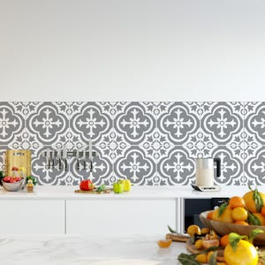 May include: A kitchen backsplash featuring square tiles with a repeating grey and white quatrefoil pattern. The wall is adorned with a magnetic knife strip holding several knives, while the white countertop displays fresh produce and a kettle.