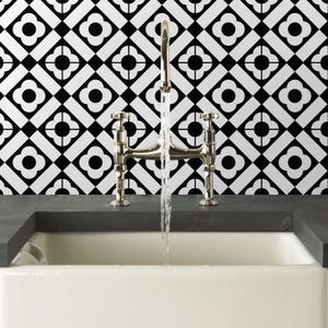Puede incluir: Un grifo de puente en cromo pulido con dos manijas que dispensa un flujo constante de agua en un fregadero blanco, frente a una pared con un patr&oacute;n de azulejos geom&eacute;tricos en blanco y negro con motivos de flores de cuatro p&eacute;talos.