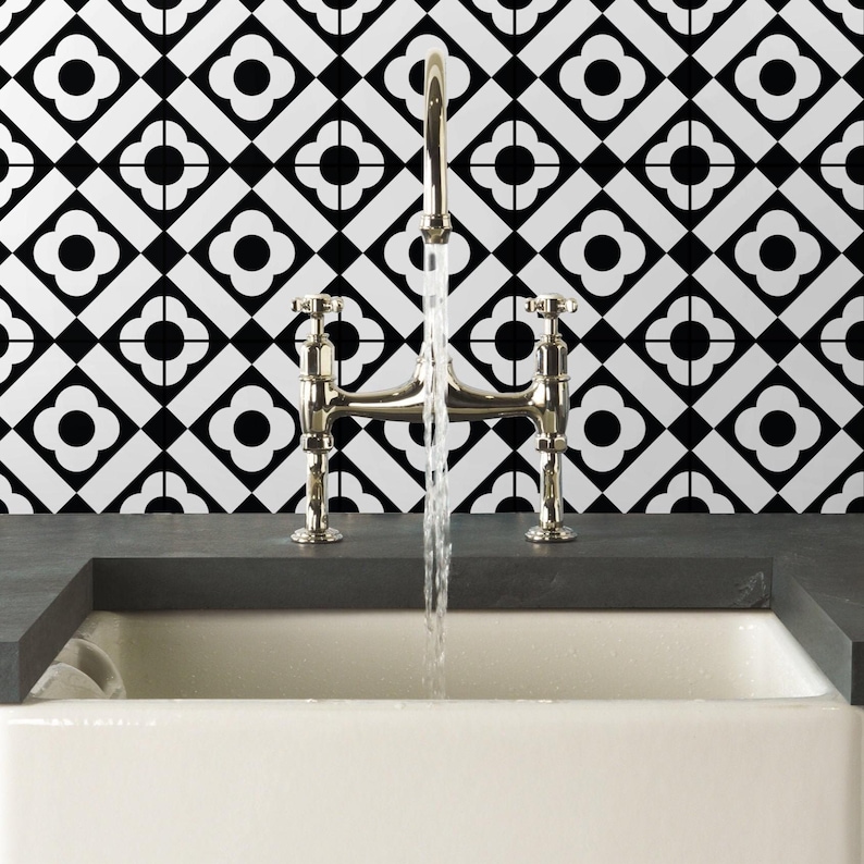 Puede incluir: Un grifo de puente en cromo pulido con dos manijas que dispensa un flujo constante de agua en un fregadero blanco, frente a una pared con un patr&oacute;n de azulejos geom&eacute;tricos en blanco y negro con motivos de flores de cuatro p&eacute;talos.