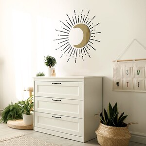 Puede incluir: Una c&oacute;moda blanca con tres cajones y tiradores negros. La c&oacute;moda est&aacute; en una habitaci&oacute;n con un espejo de media luna dorado con rayos de sol negros, una planta verde en una maceta blanca y una cesta tejida con una planta verde.