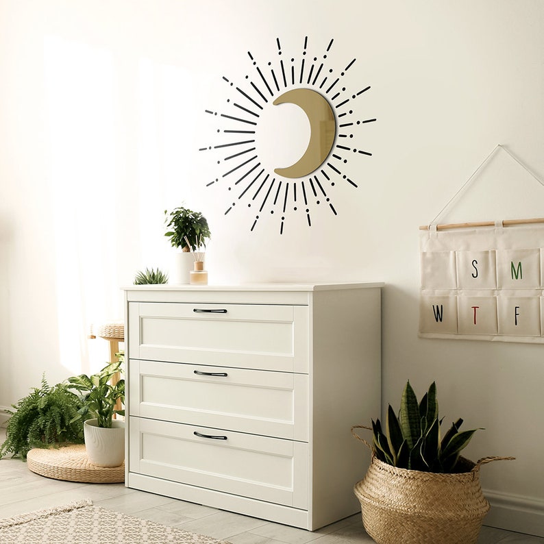 Puede incluir: Una c&oacute;moda blanca con tres cajones y tiradores negros. La c&oacute;moda est&aacute; en una habitaci&oacute;n con un espejo de media luna dorado con rayos de sol negros, una planta verde en una maceta blanca y una cesta tejida con una planta verde.