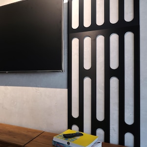 May include: A black TV remote rests on a stack of books with a yellow cover. A large black TV is mounted on a gray wall. A black decorative panel with vertical cutouts is to the right. A wooden table is in the foreground.