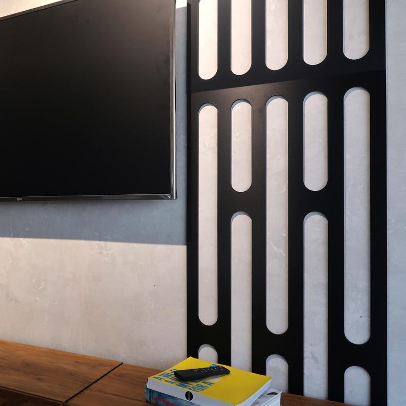 May include: A black TV remote rests on a stack of books with a yellow cover. A large black TV is mounted on a gray wall. A black decorative panel with vertical cutouts is to the right. A wooden table is in the foreground.
