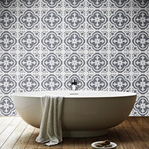 May include: A white freestanding bathtub with a white textured towel draped over the side. The wall behind the tub features a repeating pattern of dark gray and white quatrefoil tiles. The floor is made of dark brown wooden planks.