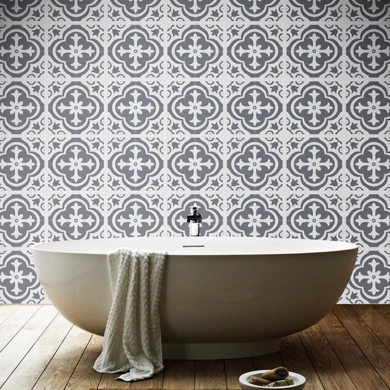 May include: A white freestanding bathtub with a white textured towel draped over the side. The wall behind the tub features a repeating pattern of dark gray and white quatrefoil tiles. The floor is made of dark brown wooden planks.