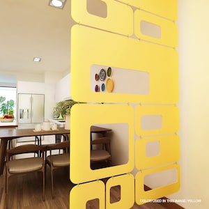 Puede incluir: Un separador de ambientes amarillo brillante con un dise&ntilde;o geom&eacute;trico. El separador est&aacute; hecho de paneles rectangulares y cuadrados conectados con ventanas recortadas. El texto "COLOR USED IN THIS IMAGE: YELLOW" es visible. Separa un comedor con mesa y sillas de madera.