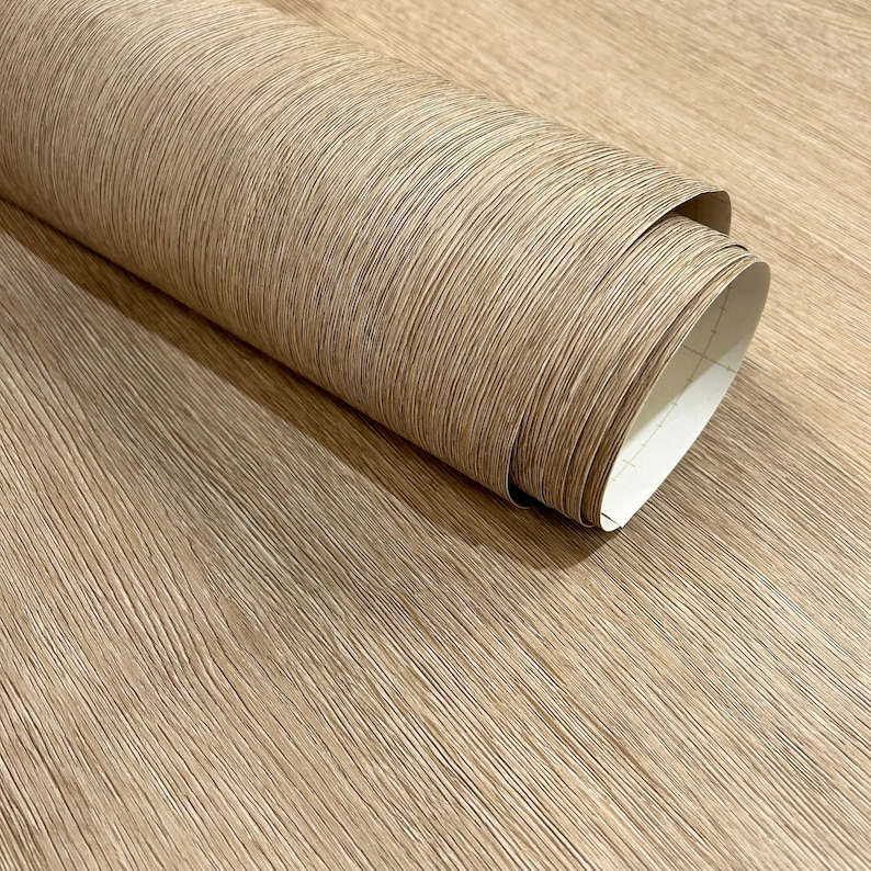 May include: A roll of light brown wood-look wallpaper with a textured, linear pattern. The wallpaper is partially unrolled, revealing the white backing. The wood grain design adds a natural aesthetic.
