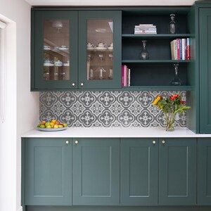 May include: Dark green kitchen cabinetry with shaker-style doors and gold hardware. The backsplash features a gray and white patterned tile design. A white countertop holds a bowl of fruit and a glass vase with a bouquet of sunflowers and roses.