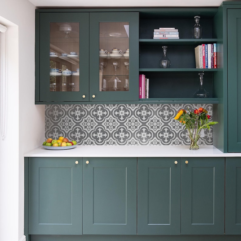 May include: Dark green kitchen cabinetry with shaker-style doors and gold hardware. The backsplash features a gray and white patterned tile design. A white countertop holds a bowl of fruit and a glass vase with a bouquet of sunflowers and roses.
