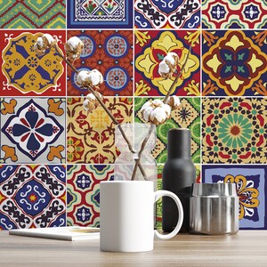 May include: A white ceramic mug sits on a wooden surface in front of a wall covered in colorful, patterned square tiles. A clear glass vase holds dried cotton stems, next to a matte black bottle and a metallic silver cylindrical container.