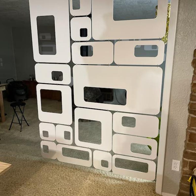 Puede incluir: Un separador de ambientes modular blanco con recortes rectangulares y cuadrados. Los paneles est&aacute;n dispuestos en un patr&oacute;n de rejilla, creando un dise&ntilde;o moderno y geom&eacute;trico. Los recortes var&iacute;an en tama&ntilde;o y forma, agregando inter&eacute;s visual al separador.