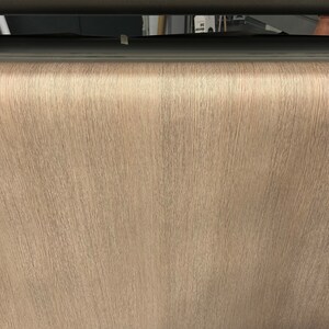 May include: A close-up of a roll of light brown textured wallpaper. The wallpaper has a vertical grain pattern, and the color is a neutral beige. The image is well-lit, showing the details of the paper's surface.