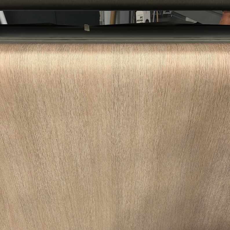 May include: A close-up of a roll of light brown textured wallpaper. The wallpaper has a vertical grain pattern, and the color is a neutral beige. The image is well-lit, showing the details of the paper's surface.