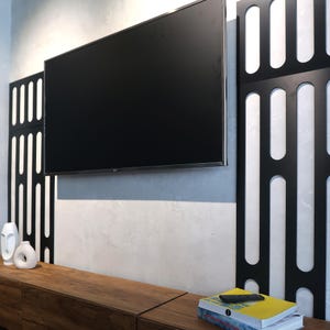 May include: A large, black flat-screen television mounted on a light gray wall. Decorative black panels with white accents flank the TV. A wooden cabinet below holds white vases and books. A remote control rests on a stack of books.