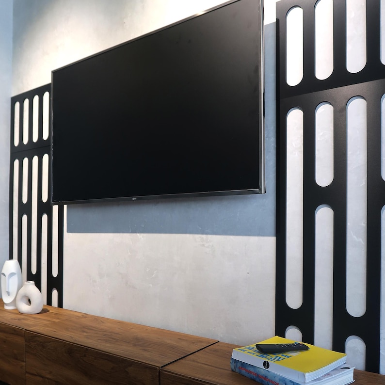May include: A large, black flat-screen television mounted on a light gray wall. Decorative black panels with white accents flank the TV. A wooden cabinet below holds white vases and books. A remote control rests on a stack of books.