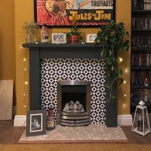 May include: A dark gray fireplace mantel featuring a black and white geometric diamond tile surround, a silver metal firebox with gray stones, and a light-colored speckled hearth. The mantel is decorated with various framed pictures, plants, and lights.