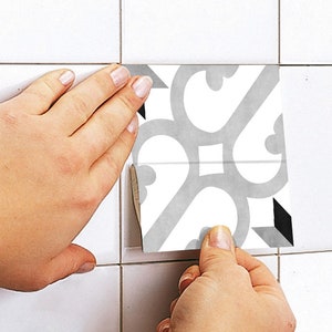 May include: A square adhesive tile decal featuring a light gray and white symmetrical scrollwork pattern with small black geometric accents, being applied to a white tiled surface to cover an existing square tile section.