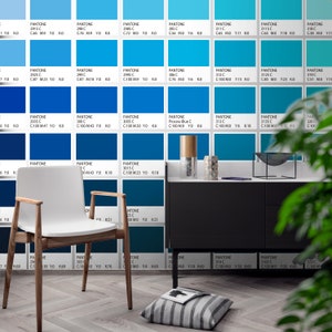 May include: A white chair with wooden legs and arms sits before a wall covered in a grid of blue Pantone color swatches, each labeled with its specific code and CMYK values. A black cabinet, a striped floor cushion, and a book are also present.