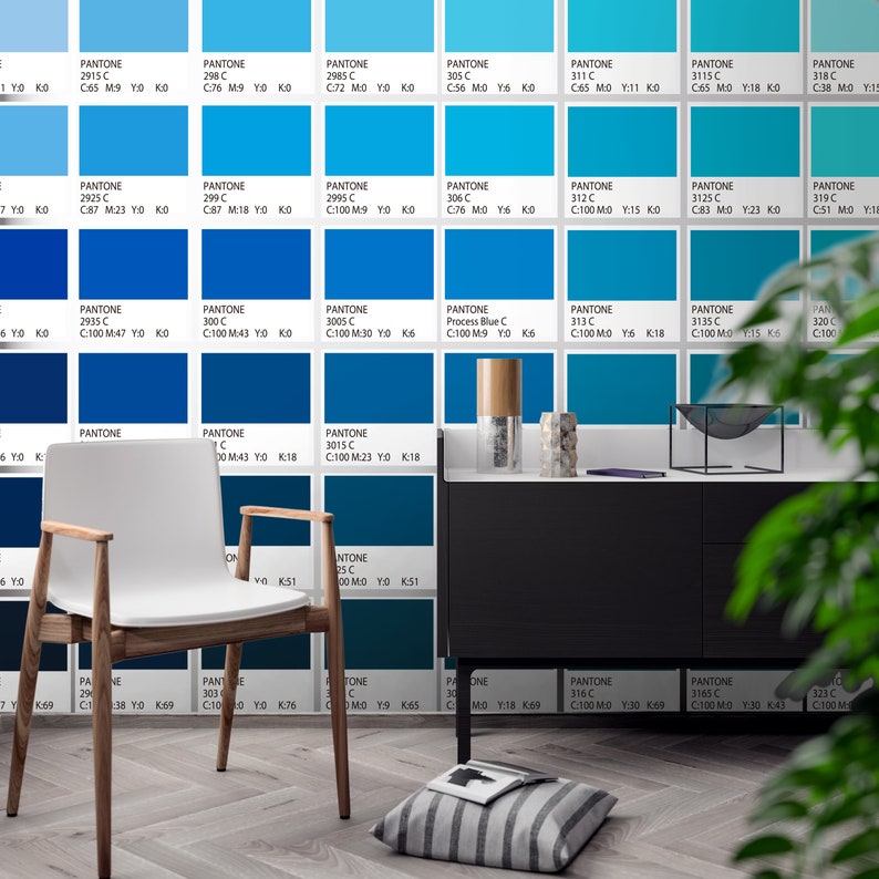 May include: A white chair with wooden legs and arms sits before a wall covered in a grid of blue Pantone color swatches, each labeled with its specific code and CMYK values. A black cabinet, a striped floor cushion, and a book are also present.