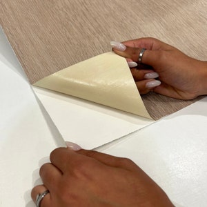 May include: A roll of light brown wood-grain contact paper being peeled back to reveal a white backing. The contact paper has a textured surface and is being held by two hands.