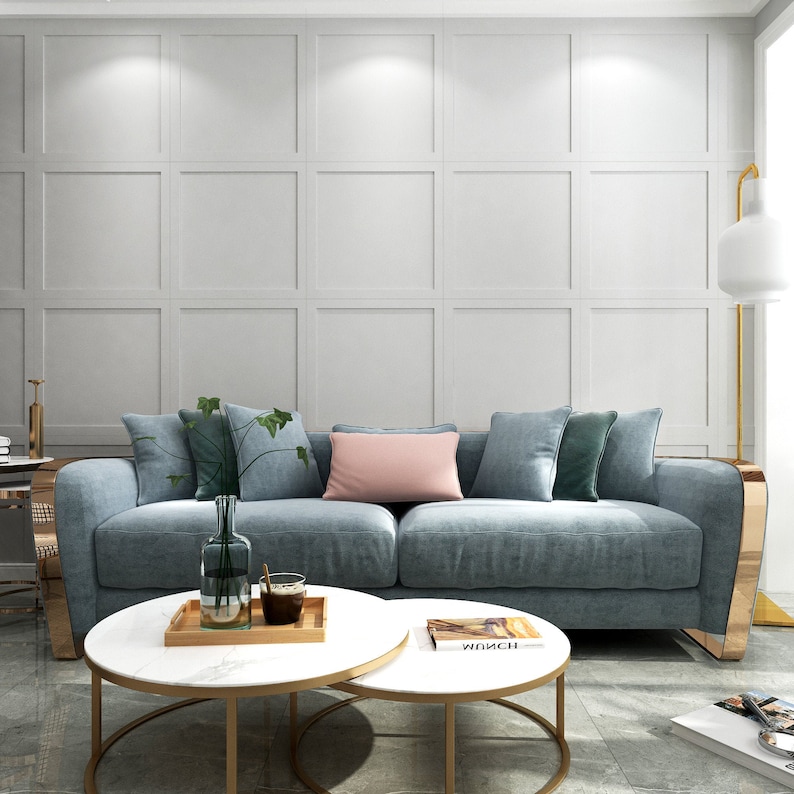 May include: A light blue sofa with gold accents sits in a living room with a white paneled wall. The sofa has several pillows, including a pink one. Two round nesting coffee tables with a marble top and gold metal frame are in front of the sofa. A book titled "Wunsch" is on the top table.