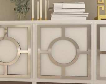 May include: A white cabinet with silver geometric door accents. The cabinet has a rectangular shape with a flat top. The doors feature a circular design within a square frame. The cabinet is decorated with candles, books, and decorative items.