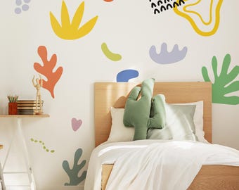 May include: A bedroom scene with a wooden bed frame, white bedding, and a green cactus-shaped pillow. The wall is decorated with colorful abstract shapes in orange, yellow, green, blue, and purple. A small desk with art supplies is visible.