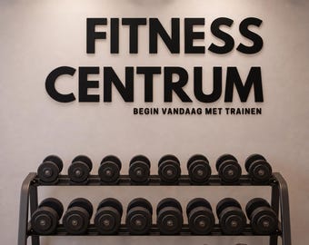 May include: A black metal two-tier dumbbell rack holding sixteen black dumbbells, with a pair of silver dumbbells on the floor. The wall features black 3D lettering that reads FITNESS CENTRUM and the smaller text BEGIN VANDAAG MET TRAINEN.