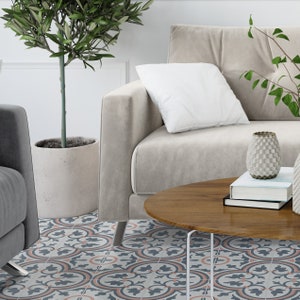 Puede incluir: Un sof&aacute; beige con un coj&iacute;n blanco, una mesa de centro redonda de madera con libros apilados y un jarr&oacute;n texturizado, y un olivo en una maceta de hormig&oacute;n, sobre un suelo con un patr&oacute;n floral repetitivo en azul, gris y melocot&oacute;n.