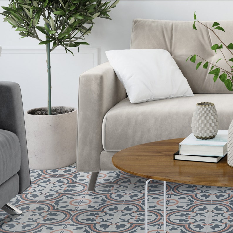 Puede incluir: Un sof&aacute; beige con un coj&iacute;n blanco, una mesa de centro redonda de madera con libros apilados y un jarr&oacute;n texturizado, y un olivo en una maceta de hormig&oacute;n, sobre un suelo con un patr&oacute;n floral repetitivo en azul, gris y melocot&oacute;n.