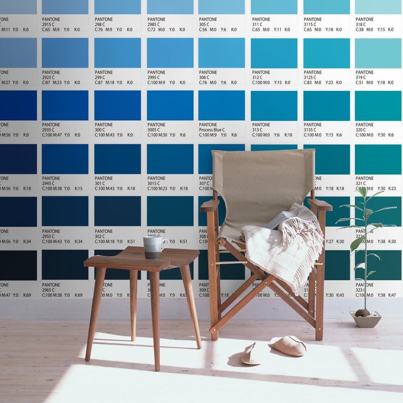 May include: A wall covered in a grid of blue Pantone color swatches with printed codes, featuring a wooden stool with a two-toned mug, a wooden folding chair with a beige fabric seat draped with a light blanket, and a small potted green plant.