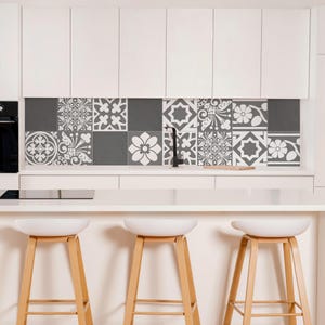 May include: A kitchen backsplash featuring a grid of square tiles with alternating dark gray solid colors and white floral or geometric patterns. The tiles are positioned between white upper cabinets and a white countertop with three stools.