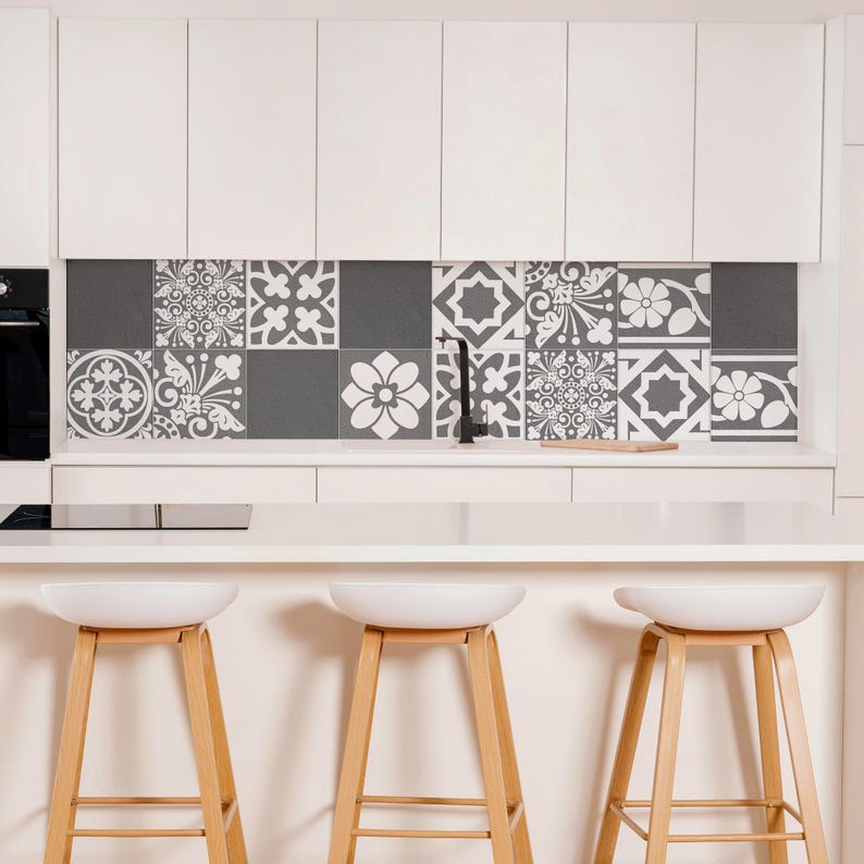 May include: A kitchen backsplash featuring a grid of square tiles with alternating dark gray solid colors and white floral or geometric patterns. The tiles are positioned between white upper cabinets and a white countertop with three stools.