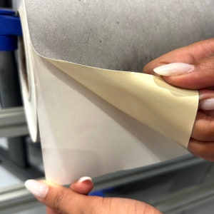 K&ouml;nnte beinhalten: Eine Rolle aus wei&szlig;em und beigem Material wird teilweise abgerollt und zeigt eine beige Unterseite. Das Material scheint eine Art selbstklebendes Vinyl oder Papier mit glatter, matter Oberfl&auml;che zu sein. Die Kanten sind sauber und gerade.