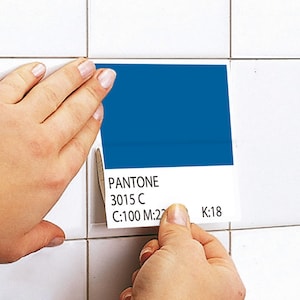 May include: A square Pantone color swatch card featuring a solid deep blue block at the top and the text PANTONE 3015 C, C:100 M:22 K:18 printed in black at the bottom, held against a white tiled surface to compare the color match.