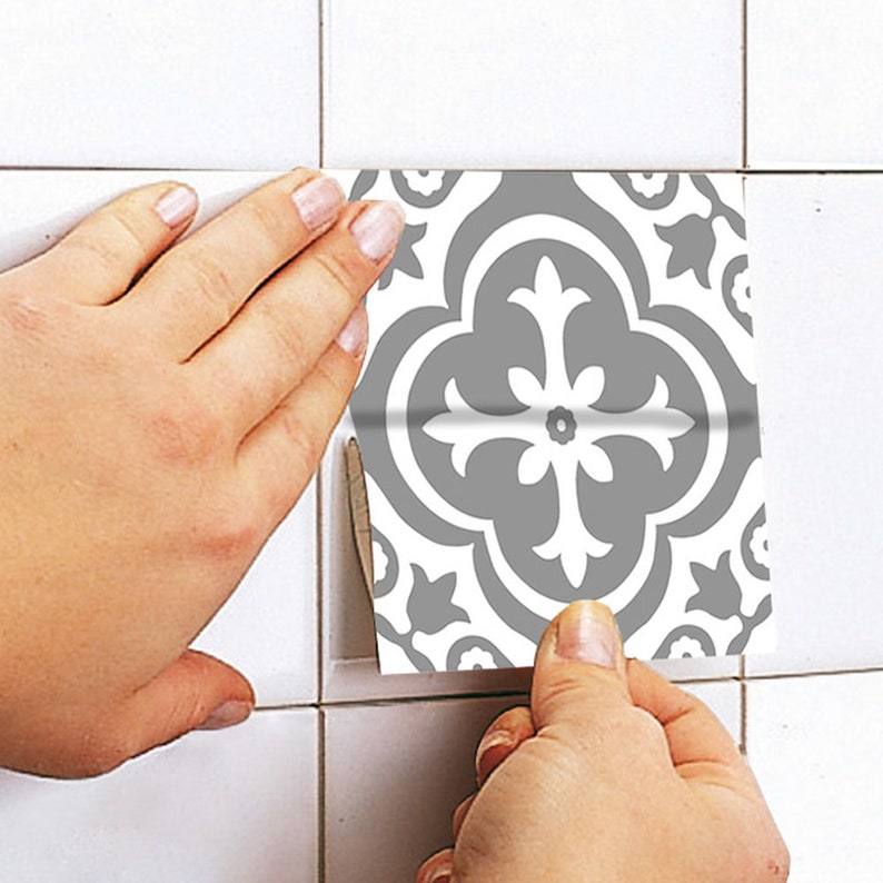 May include: A square adhesive tile decal featuring a grey and white quatrefoil pattern. The decal is being applied to a white tiled surface, showing a symmetrical floral-inspired design with curved lines and pointed edges in a repeating motif.