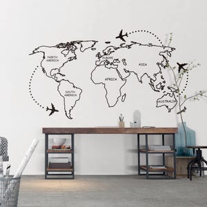 K&ouml;nnte beinhalten: Schwarzer Weltkarten-Wandtattoo mit Flugzeug-Illustrationen und gepunkteten Flugrouten. Die Karte zeigt die Kontinente Nordamerika, S&uuml;damerika, Europa, Afrika, Asien und Australien. Auf einer wei&szlig;en Wand angebracht.